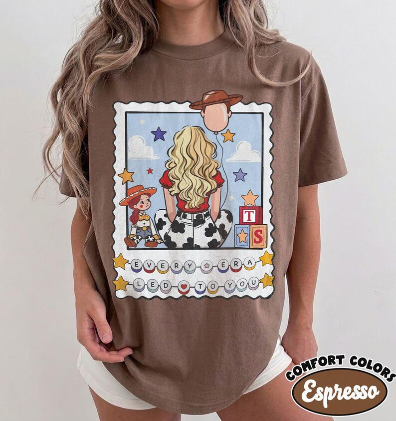 May include: A brown t-shirt featuring a graphic print of a person with long blonde hair, a cowgirl doll, and a cowboy hat balloon. The design includes alphabet blocks, stars, clouds, and the text "EVERY ERA LED TO YOU" on circular beads.