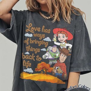 May include: A charcoal gray oversized t-shirt featuring a graphic print of Jessie, Buzz Lightyear, and Woody from Toy Story. The text reads, Love has ways of bringing things back to life, accompanied by a tree and rolling hills. The shirt is labeled Comfort Colors Pepper.