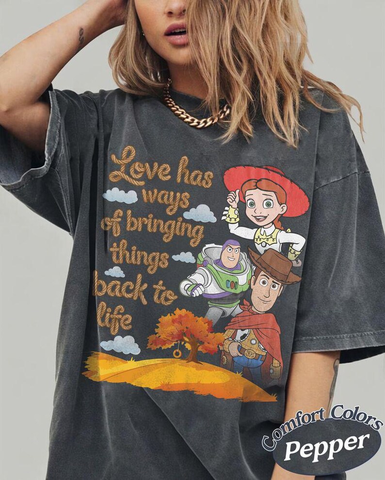May include: A charcoal gray oversized t-shirt featuring a graphic print of Jessie, Buzz Lightyear, and Woody from Toy Story. The text reads, Love has ways of bringing things back to life, accompanied by a tree and rolling hills. The shirt is labeled Comfort Colors Pepper.