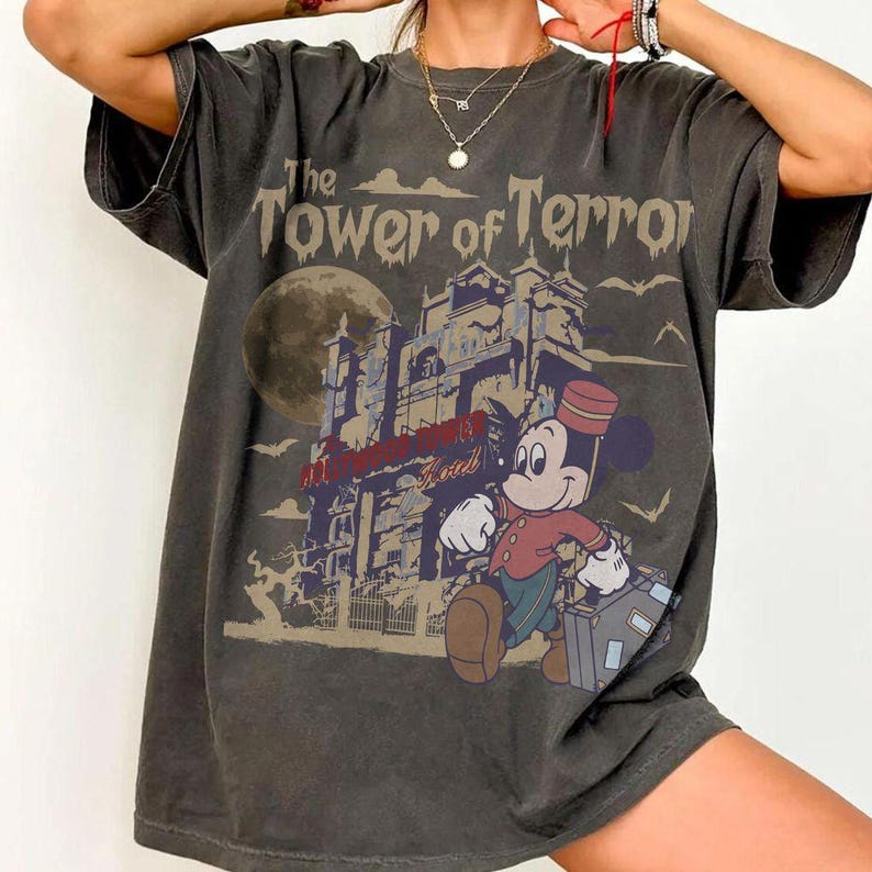 Puede incluir: Camiseta gris oscuro con un gr&aacute;fico de Mickey Mouse como botones, llevando una maleta frente a un hotel embrujado. El texto "The Tower of Terror" est&aacute; encima de la imagen, con una luna llena en el fondo.