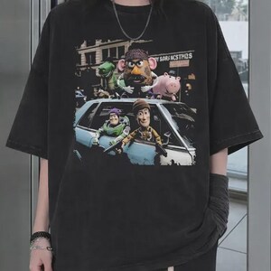 Puede incluir: Una camiseta negra de manga corta con un estampado gr&aacute;fico de cinco personajes animados de la franquicia Toy Story. El dise&ntilde;o muestra a los personajes posicionados dentro y alrededor de un coche azul claro, con un fondo de calle urbana.