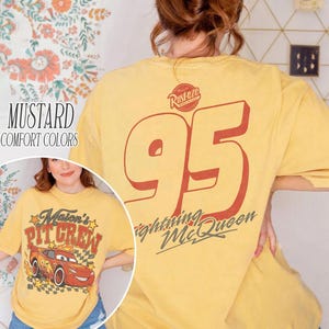Op de afbeelding: Een mosterdgeel t-shirt met een groot rood nummer 95 en de tekst Rusteze en Lightning McQueen op de achterkant. De afbeelding op de voorkant toont een rode raceauto met het nummer 95, geblokte patronen, sterren en de tekst Mason's Pit Crew.