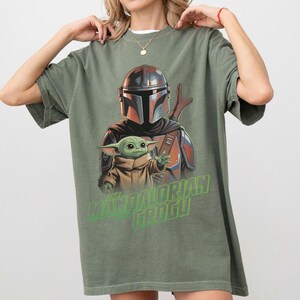 May include: An olive green short-sleeve t-shirt featuring a graphic print of The Mandalorian and Grogu. The design shows the armored character standing behind the small green creature. The text THE MANDALORIAN AND GROGU is printed in bright green.