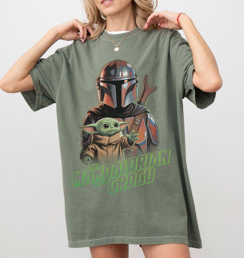 May include: An olive green short-sleeve t-shirt featuring a graphic print of The Mandalorian and Grogu. The design shows the armored character standing behind the small green creature. The text THE MANDALORIAN AND GROGU is printed in bright green.