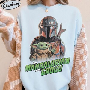 May include: A light blue crewneck sweatshirt featuring a graphic print of The Mandalorian and Grogu. The design shows the armored Mandalorian character standing behind the small green Grogu character. The text reads The Mandalorian and Grogu.