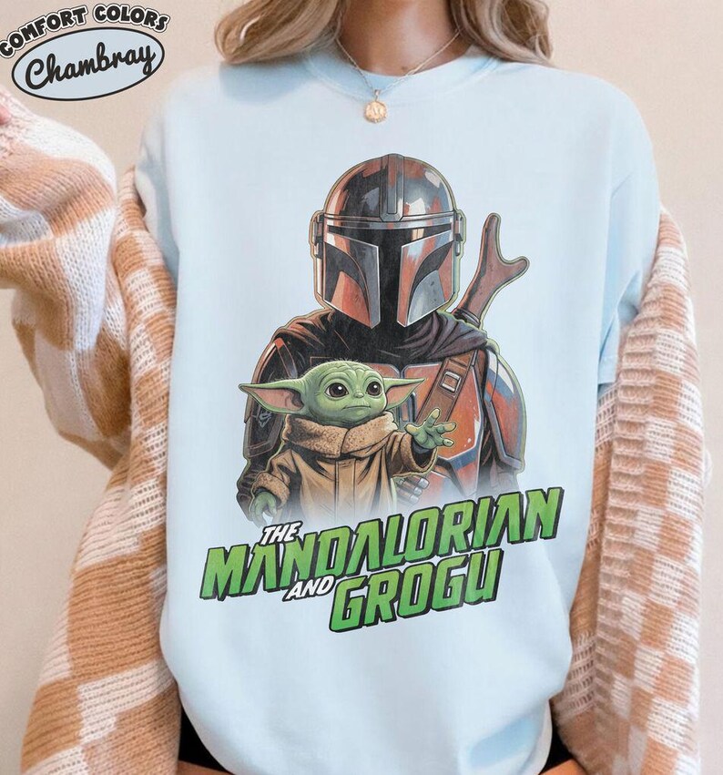 May include: A light blue crewneck sweatshirt featuring a graphic print of The Mandalorian and Grogu. The design shows the armored Mandalorian character standing behind the small green Grogu character. The text reads The Mandalorian and Grogu.