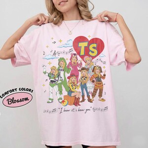Puede incluir: Una camiseta rosa claro con un gran coraz&oacute;n rojo que contiene las letras TS. Debajo del coraz&oacute;n hay siete personajes de dibujos animados con varios disfraces, rodeados de notas musicales y nubes. El texto dice "I knew it, I knew you".