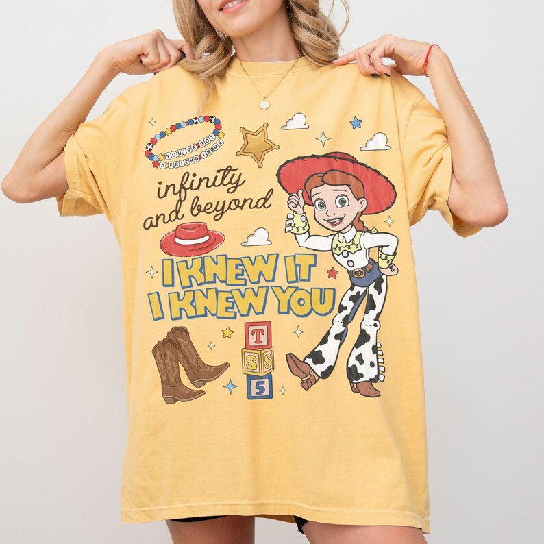 May include: A yellow short-sleeve t-shirt featuring a graphic print of a cowgirl character, a red hat, brown cowboy boots, and alphabet blocks. The text reads infinity and beyond, I KNEW IT, I KNEW YOU, and YOU'VE GOT A FRIEND IN ME on a beaded bracelet.