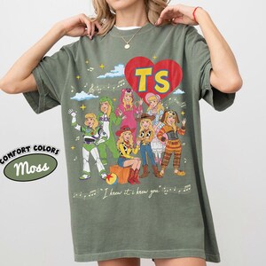 Puede incluir: Una camiseta verde musgo con un gr&aacute;fico de un gran coraz&oacute;n rojo con las letras amarillas TS en su interior. El dise&ntilde;o incluye siete personajes ilustrados con varios disfraces, notas musicales, estrellas y el texto I knew it, i knew you.