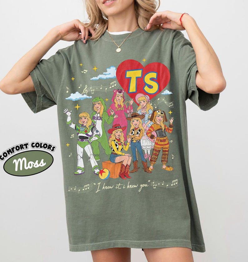 Puede incluir: Una camiseta verde musgo con un gr&aacute;fico de un gran coraz&oacute;n rojo con las letras amarillas TS en su interior. El dise&ntilde;o incluye siete personajes ilustrados con varios disfraces, notas musicales, estrellas y el texto I knew it, i knew you.