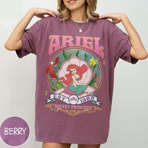 May include: Berry-coloured t-shirt with a graphic of Ariel from The Little Mermaid. The design includes the word "ARIEL" in pink, the text "EST. 1989 DISNEY PRINCESS", and illustrations of characters from the movie. The shirt has a relaxed fit.
