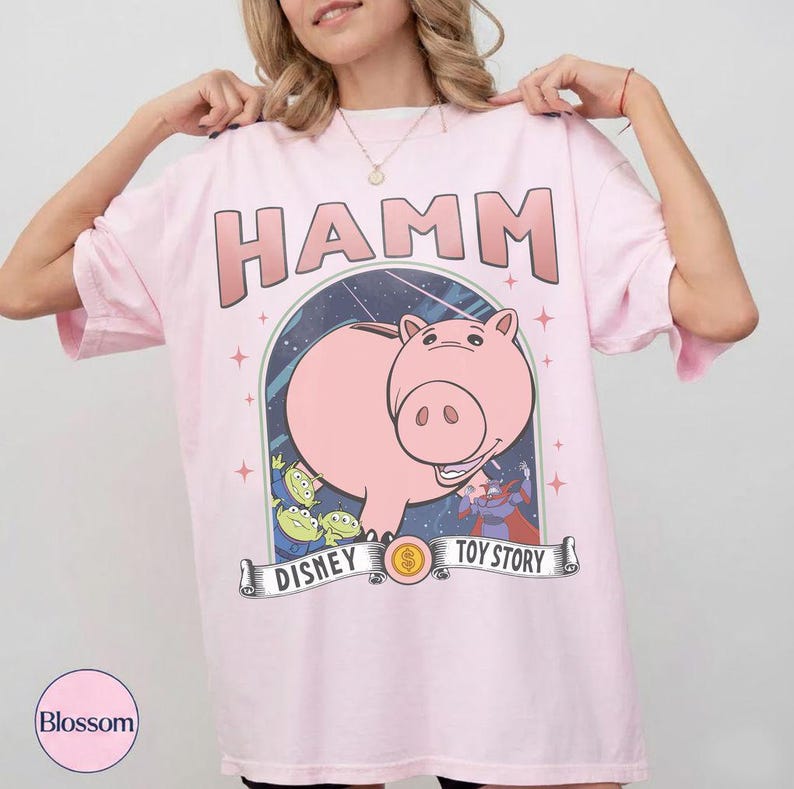 May include: Pale pink t-shirt with the word "HAMM" in a light brown font above a cartoon image of Hamm the pig from Toy Story. The design includes the text "DISNEY TOY STORY" on a banner.