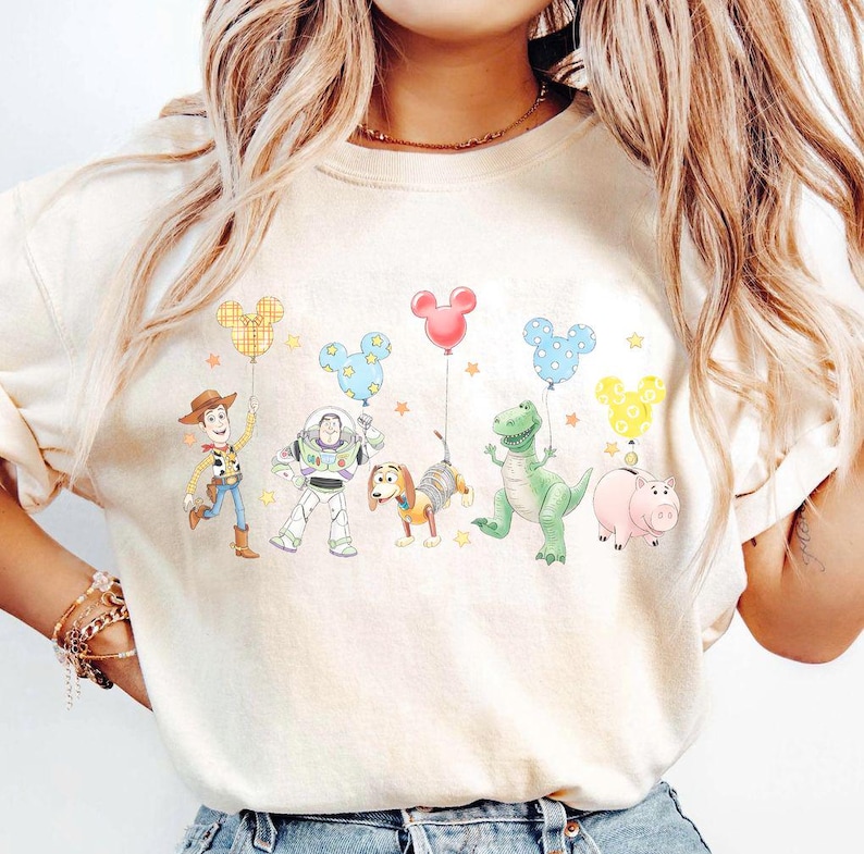 May include: Cream-coloured sweatshirt with a vibrant graphic of animated film characters. The design showcases a cowboy, space ranger, dog, dinosaur, and pig, each holding a Mickey Mouse-shaped balloon. The design is set against a backdrop of stars.