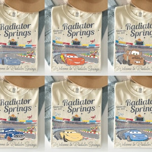 Puede incluir: Camisetas color crema con un gr&aacute;fico de una pista de carreras y el texto "Radiator Springs California est. 2012" y "Welcome to Radiator Springs." Cada camiseta muestra un coche de dibujos animados diferente. Un sombrero marr&oacute;n y mezclilla est&aacute;n en el fondo.