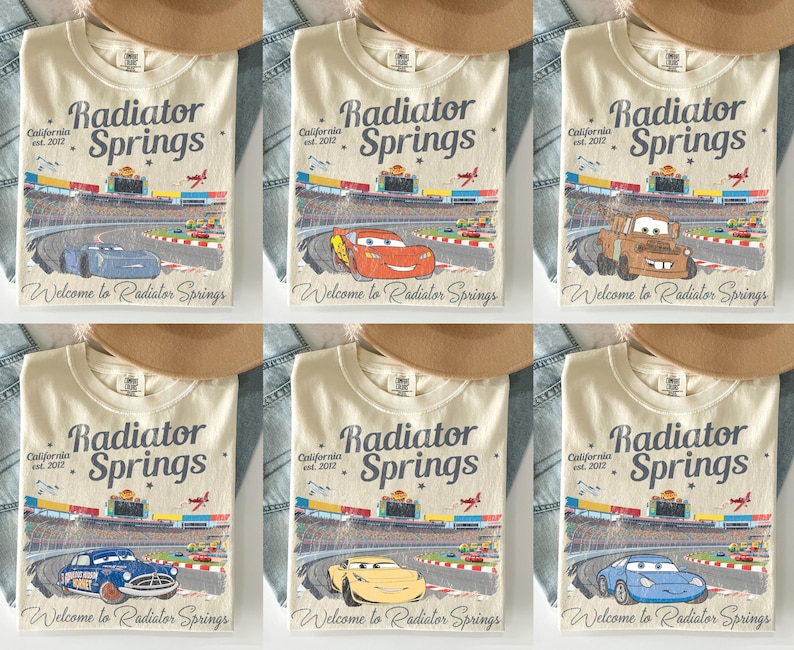 Puede incluir: Camisetas color crema con un gr&aacute;fico de una pista de carreras y el texto "Radiator Springs California est. 2012" y "Welcome to Radiator Springs." Cada camiseta muestra un coche de dibujos animados diferente. Un sombrero marr&oacute;n y mezclilla est&aacute;n en el fondo.