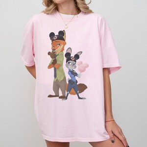 Pu&ograve; includere: T-shirt rosa chiaro con un'illustrazione a cartoni animati di Nick Wilde e Judy Hopps di Zootropolis. Nick, una volpe, indossa una camicia verde e Judy, una coniglia, &egrave; in uniforme da poliziotta. Entrambi i personaggi indossano orecchie di Topolino.