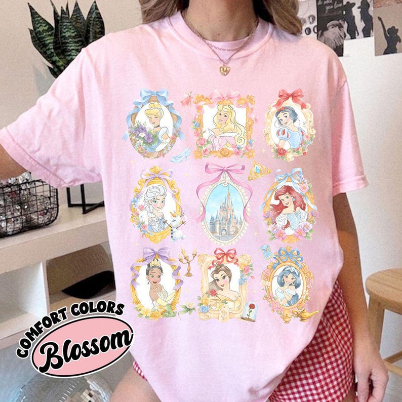 May include: Pink t-shirt featuring nine framed portraits of animated princesses. Each portrait is surrounded by floral and bow accents. The shirt has the words "COMFORT COLORS" and "Blossom" printed on the front.