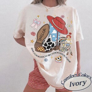 May include: An ivory t-shirt featuring a graphic print of a cowboy boot with a cow print pattern, a red cowboy hat, a small character figure, and stacked blocks with the letters BTS. The text TAYLOR and I KNEW IT I KNEW YOU are included.