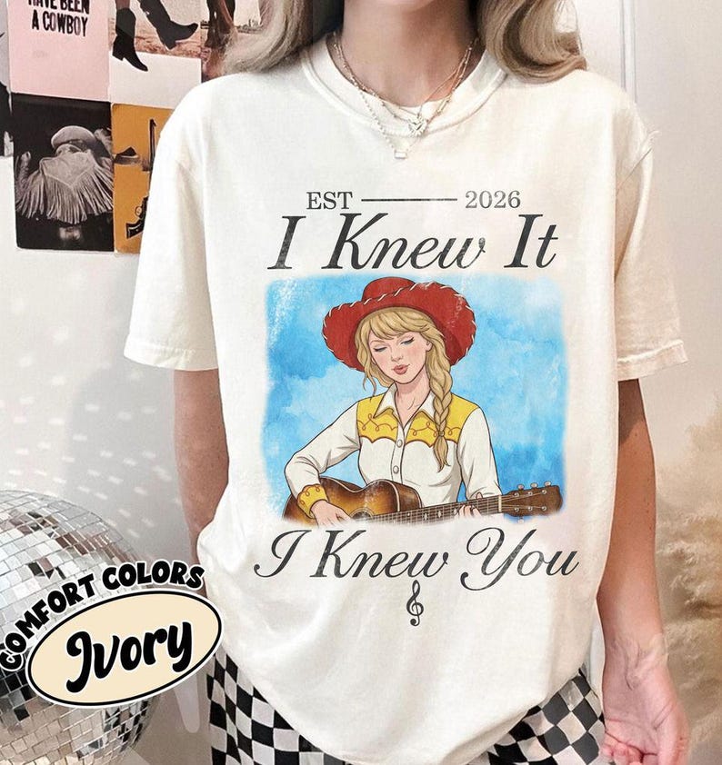 May include: An ivory short-sleeve t-shirt featuring a graphic of a person with blonde hair wearing a red cowboy hat and a yellow and white shirt while playing an acoustic guitar. The text reads EST 2026 I Knew It I Knew You with a music note.