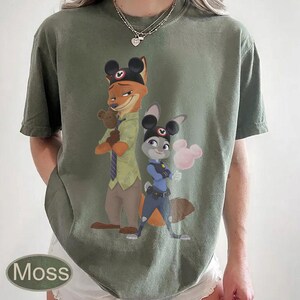 Pu&ograve; includere: T-shirt verde muschio con un'illustrazione a cartoni animati di Nick Wilde e Judy Hopps di Zootropolis, con orecchie di Topolino. Nick tiene un orsacchiotto e Judy uno zucchero filato rosa. La parola "Moss" &egrave; stampata in basso a sinistra.