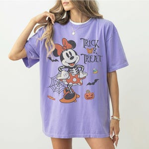 May include: A light purple short-sleeve t-shirt featuring a graphic of a cartoon mouse dressed in a skeleton costume with a red bow. The design includes the text Trick R Treat, a spider web, bats, a jack-o'-lantern, and candy corn icons.