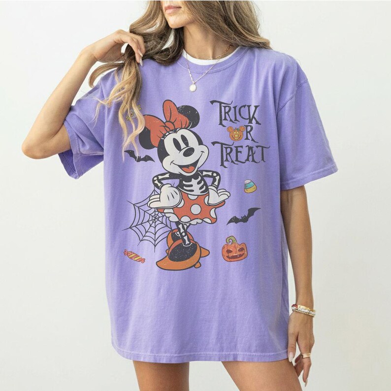 May include: A light purple short-sleeve t-shirt featuring a graphic of a cartoon mouse dressed in a skeleton costume with a red bow. The design includes the text Trick R Treat, a spider web, bats, a jack-o'-lantern, and candy corn icons.