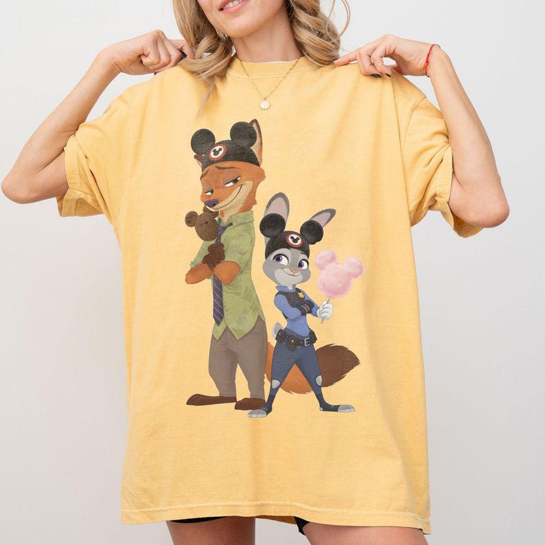 Pu&ograve; includere: T-shirt giallo con un'illustrazione di Nick Wilde e Judy Hopps di Zootropolis, con orecchie di Topolino. Nick tiene un orsacchiotto e Judy tiene lo zucchero filato. La maglietta &egrave; in stile casual.