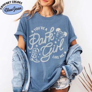 Op de afbeelding: Een blauw T-shirt met korte mouwen en de witte tekst "YOU'RE A Park Girl LIKE ME" in een touwachtig lettertype. Het ontwerp bevat een afbeelding van een cowboylaars, muizenoortjes en kleine sterretjes. Een spijkerjasje hangt over de schouders.