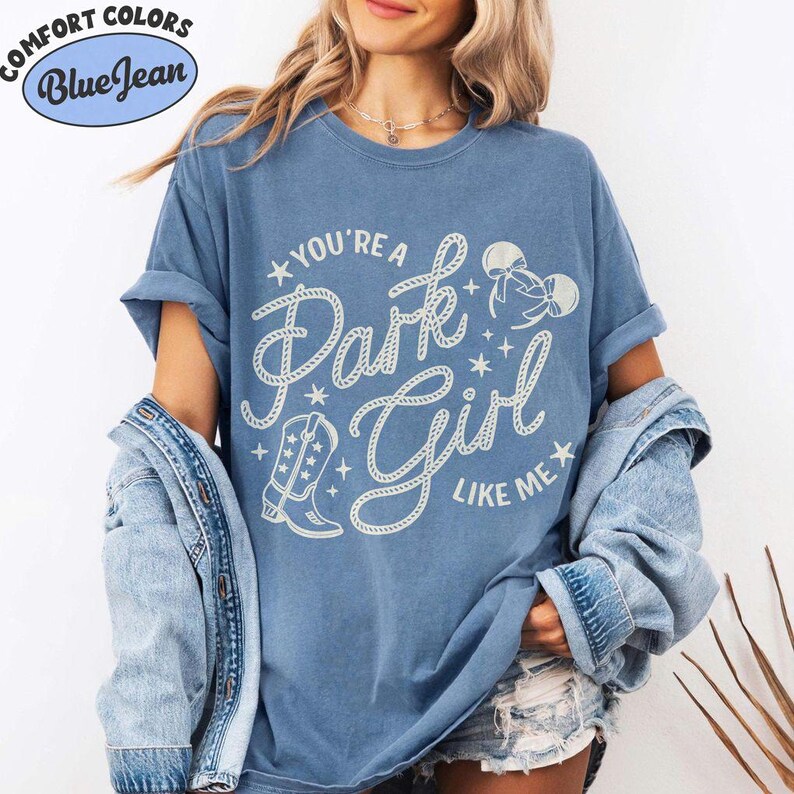 Op de afbeelding: Een blauw T-shirt met korte mouwen en de witte tekst "YOU'RE A Park Girl LIKE ME" in een touwachtig lettertype. Het ontwerp bevat een afbeelding van een cowboylaars, muizenoortjes en kleine sterretjes. Een spijkerjasje hangt over de schouders.
