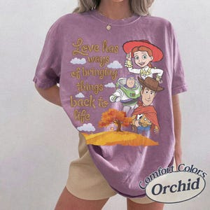 May include: A purple short-sleeve t-shirt featuring a graphic of Jessie, Buzz Lightyear, and Woody from Toy Story. The text reads, Love has ways of bringing things back to life, alongside a tree and clouds. The shirt has a crew neckline.