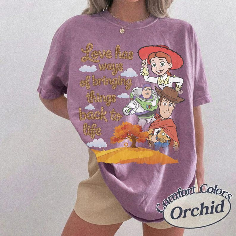 May include: A purple short-sleeve t-shirt featuring a graphic of Jessie, Buzz Lightyear, and Woody from Toy Story. The text reads, Love has ways of bringing things back to life, alongside a tree and clouds. The shirt has a crew neckline.