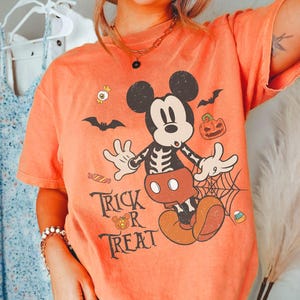 May include: An orange t-shirt featuring a graphic of Mickey Mouse with a skeleton ribcage design. The print includes bats, a jack-o'-lantern, candy, a spiderweb, and the text "TRICK OR TREAT" in a black serif font. The shirt has short sleeves.