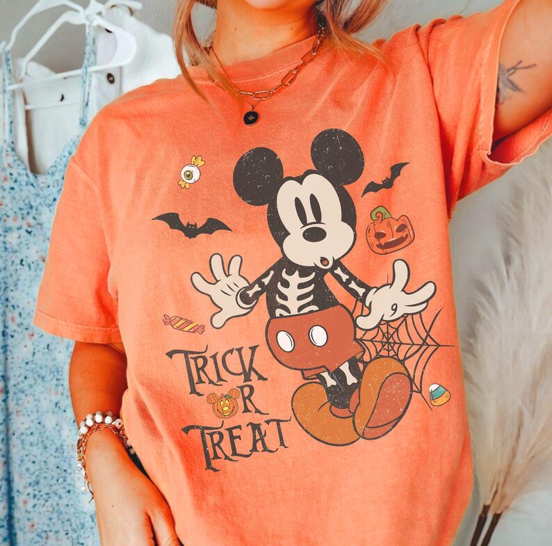 May include: An orange t-shirt featuring a graphic of Mickey Mouse with a skeleton ribcage design. The print includes bats, a jack-o'-lantern, candy, a spiderweb, and the text "TRICK OR TREAT" in a black serif font. The shirt has short sleeves.