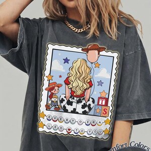 May include: A charcoal grey t-shirt featuring a graphic print of a blonde character with a red shirt and cow-print pants, a small cowgirl doll, toy blocks, and a cowboy hat balloon. The text reads EVERY ERA LED TO YOU with colorful circular beads.