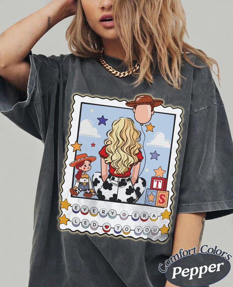 May include: A charcoal grey t-shirt featuring a graphic print of a blonde character with a red shirt and cow-print pants, a small cowgirl doll, toy blocks, and a cowboy hat balloon. The text reads EVERY ERA LED TO YOU with colorful circular beads.