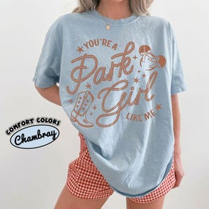 Op de afbeelding: Een lichtblauw t-shirt met korte mouwen en bruine grafische tekst die luidt YOU'RE A Park Girl LIKE ME. Het ontwerp bevat een cowboylaars, sterren en muizenoortjes. Een cirkelvormig logo in de hoek toont de tekst COMFORT COLORS Chambray.