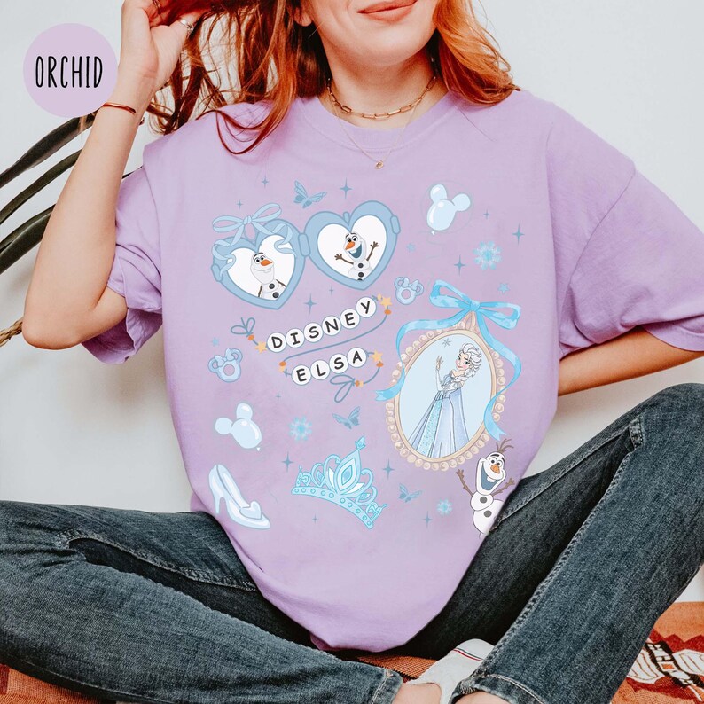May include: A light purple t-shirt featuring a Disney Elsa design. The design includes images of Elsa, Olaf, and other Frozen-themed elements. The text "DISNEY ELSA" is also visible. The shirt is a relaxed fit.