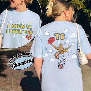 May include: A light blue t-shirt featuring a graphic on the front that reads I KNEW IT, I KNEW YOU with a heart and TS. The back graphic shows a person with a guitar, a red hat, and the text TS against a background of clouds and stars.