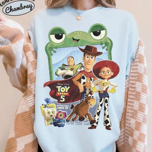 May include: A light blue crewneck sweatshirt featuring a graphic print of Toy Story 5 characters including Woody, Buzz Lightyear, Jessie, and Bullseye. The design includes a green frog frame, the Toy Story 5 logo, and the text In Theaters June 19.