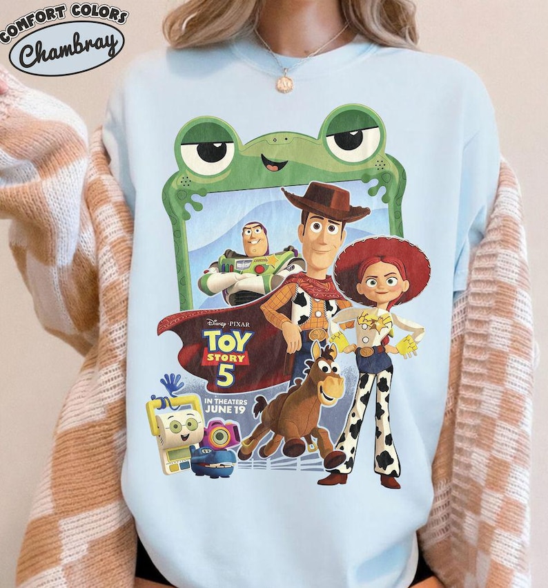May include: A light blue crewneck sweatshirt featuring a graphic print of Toy Story 5 characters including Woody, Buzz Lightyear, Jessie, and Bullseye. The design includes a green frog frame, the Toy Story 5 logo, and the text In Theaters June 19.