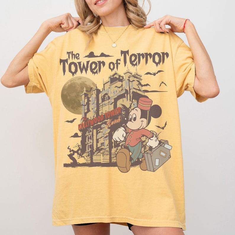 Puede incluir: Camiseta amarilla mostaza con un gr&aacute;fico de Mickey Mouse como botones frente a la Torre del Terror. El dise&ntilde;o incluye el texto "The Tower of Terror" y "Hollywood Tower Hotel". Murci&eacute;lagos y una luna llena tambi&eacute;n est&aacute;n en el dise&ntilde;o.