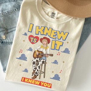 May include: A cream-colored t-shirt featuring a graphic of a person in a cowboy hat playing an acoustic guitar while sitting on a stool. The text reads I KNEW TS IT and I KNEW YOU, surrounded by blue clouds, red musical notes, and gold stars.
