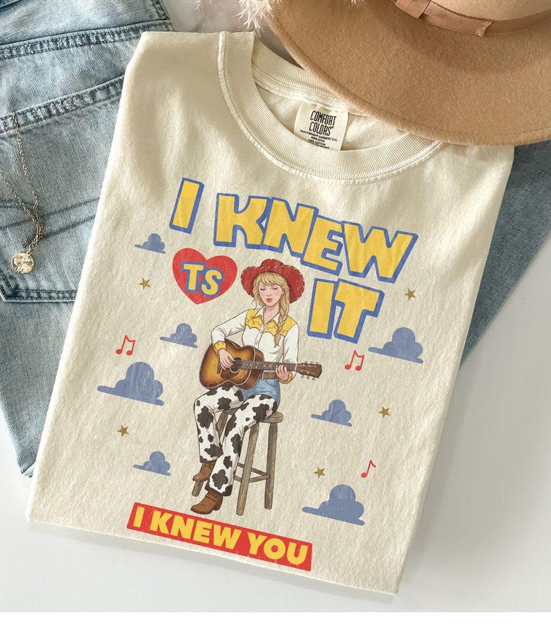 May include: A cream-colored t-shirt featuring a graphic of a person in a cowboy hat playing an acoustic guitar while sitting on a stool. The text reads I KNEW TS IT and I KNEW YOU, surrounded by blue clouds, red musical notes, and gold stars.