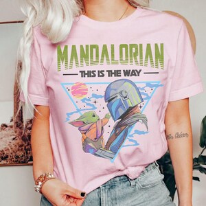 Może przedstawiać: Jasnor&oacute;żowy t-shirt z nadrukiem graficznym przedstawiającym Mandalorianina i Grogu. Napis "MANDALORIAN" widnieje w kolorze zielonym nad frazą "THIS IS THE WAY" w kolorze czarnym. Wz&oacute;r zawiera niebieski tr&oacute;jkąt, r&oacute;żową planetę i gwiazdy.