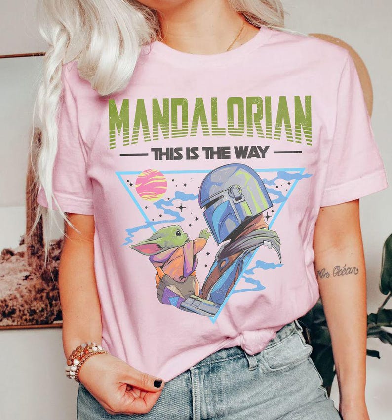 Może przedstawiać: Jasnor&oacute;żowy t-shirt z nadrukiem graficznym przedstawiającym Mandalorianina i Grogu. Napis "MANDALORIAN" widnieje w kolorze zielonym nad frazą "THIS IS THE WAY" w kolorze czarnym. Wz&oacute;r zawiera niebieski tr&oacute;jkąt, r&oacute;żową planetę i gwiazdy.