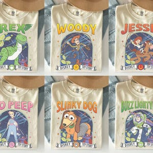 May include: Six cream-coloured t-shirts featuring characters from Disney's Toy Story. Each shirt displays a different character: Rex, Woody, Jessie, Bo Peep, Slinky Dog, and Buzz Lightyear, with the Disney Toy Story logo.