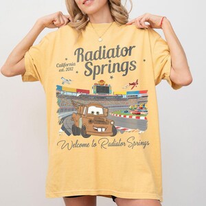 Puede incluir: Camiseta de color amarillo mostaza con un gr&aacute;fico de Mate de la pel&iacute;cula Cars en una pista de carreras. La camiseta tiene el texto "Radiator Springs" y "Welcome to Radiator Springs". La camiseta tambi&eacute;n tiene el texto "California est. 2012".