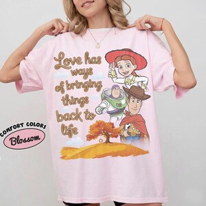 May include: A light pink t-shirt featuring a graphic print of Jessie, Buzz Lightyear, and Woody from Toy Story. The text reads, Love has ways of bringing things back to life. Below the characters is an orange tree on a hill. A Comfort Colors logo is visible.