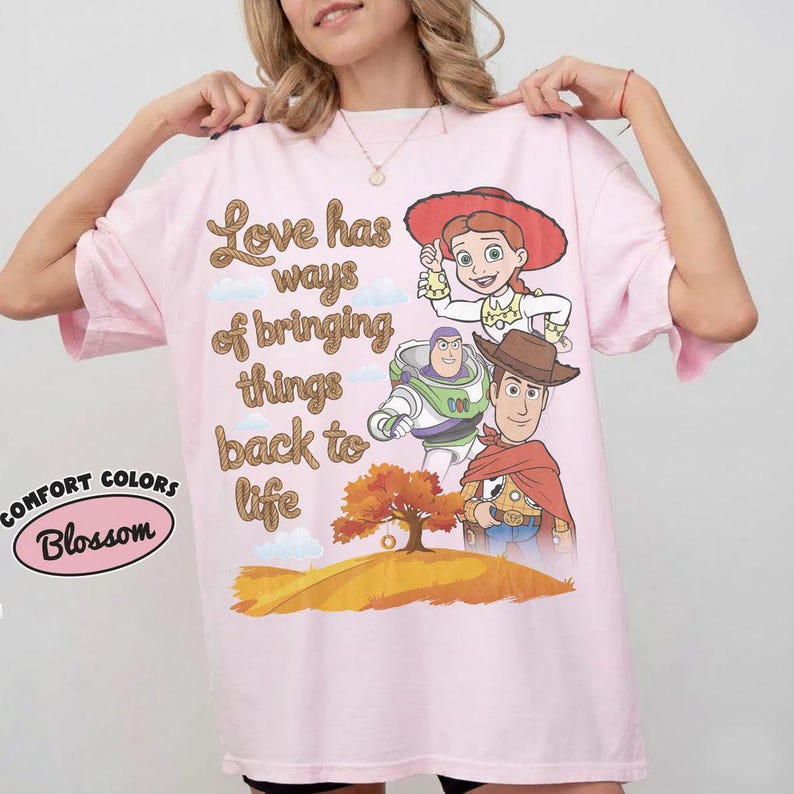 May include: A light pink t-shirt featuring a graphic print of Jessie, Buzz Lightyear, and Woody from Toy Story. The text reads, Love has ways of bringing things back to life. Below the characters is an orange tree on a hill. A Comfort Colors logo is visible.