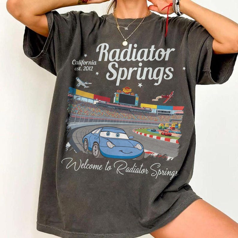 Puede incluir: Camiseta gris oscuro con un gr&aacute;fico de un coche de dibujos animados azul en un circuito. El texto dice "Radiator Springs", "Welcome to Radiator Springs" y "California est. 2012". El dise&ntilde;o est&aacute; sobre un fondo de circuito.